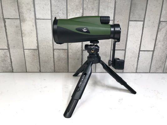 Green and black spotting scope on a tripod against a tiled wall.