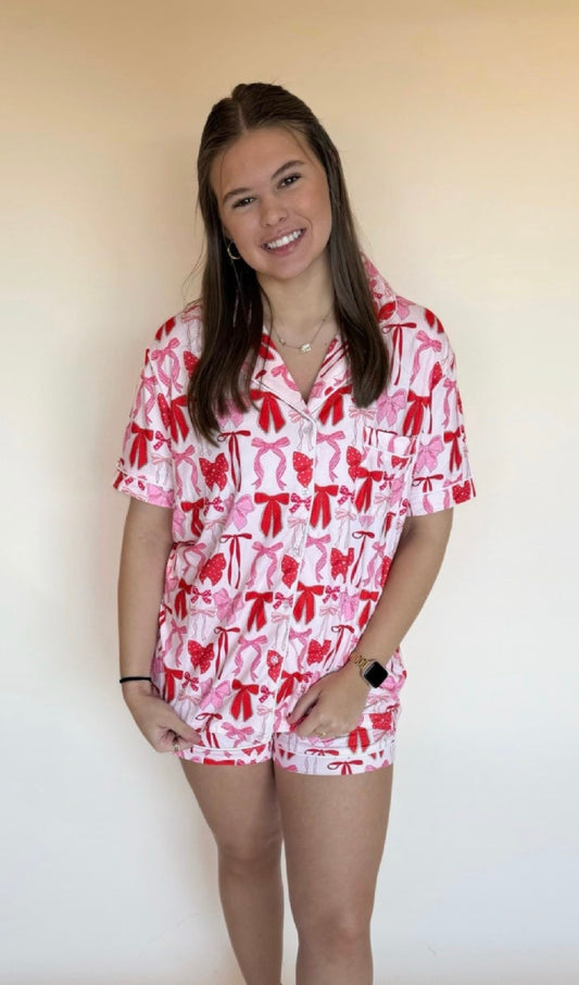Woman wearing a pink short-sleeve pajama set with red and pink bow print and matching shorts, standing against a neutral beige backdrop. 