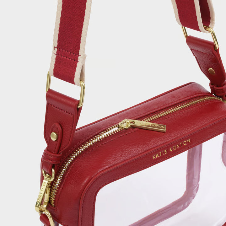 A red leather crossbody bag with a clear front window, gold-tone hardware, and a red-and-cream striped shoulder strap. The brand name &