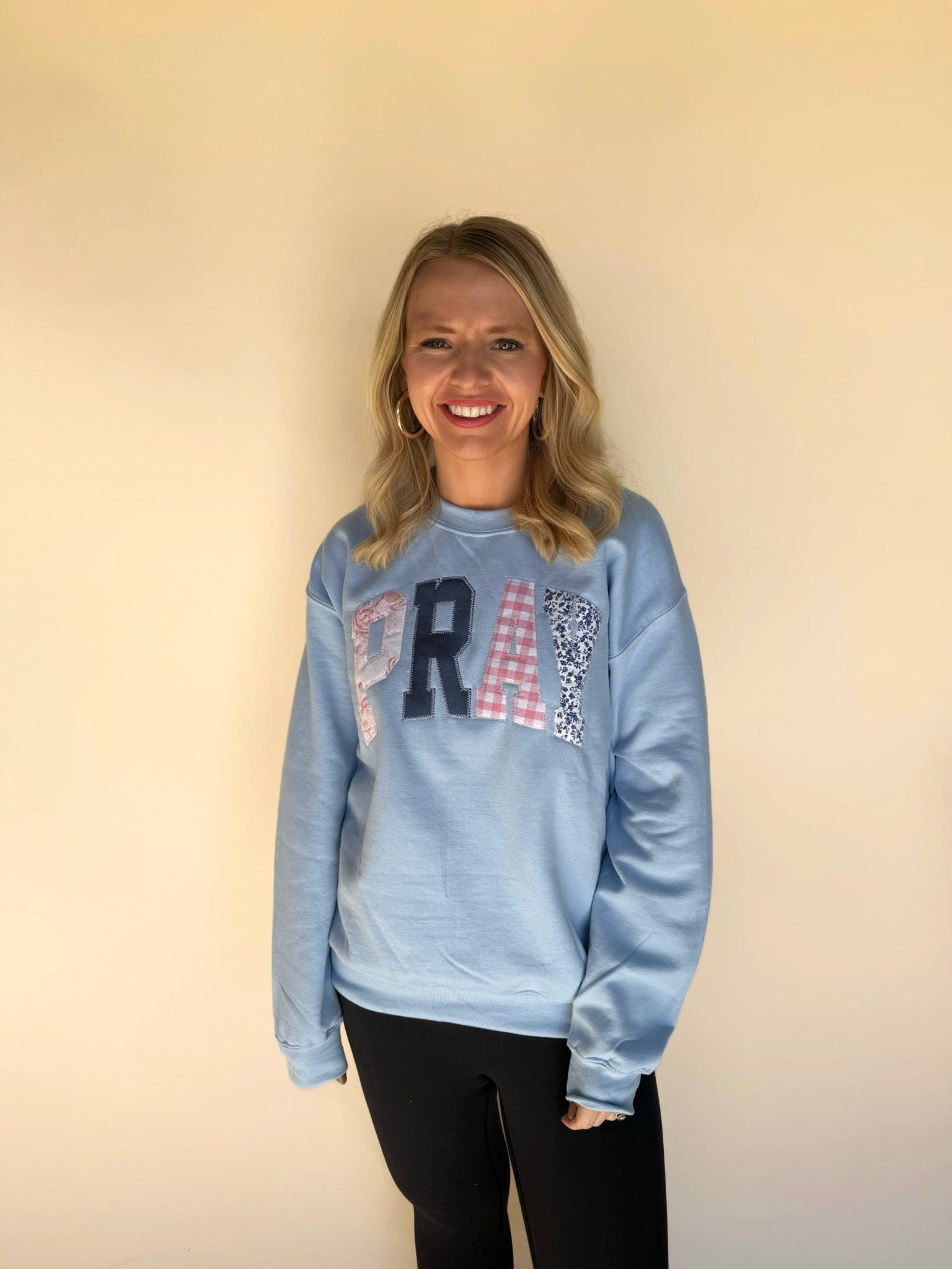 Woman wearing a light blue sweatshirt with appliqué letters spelling “PRAY,” paired with black leggings.