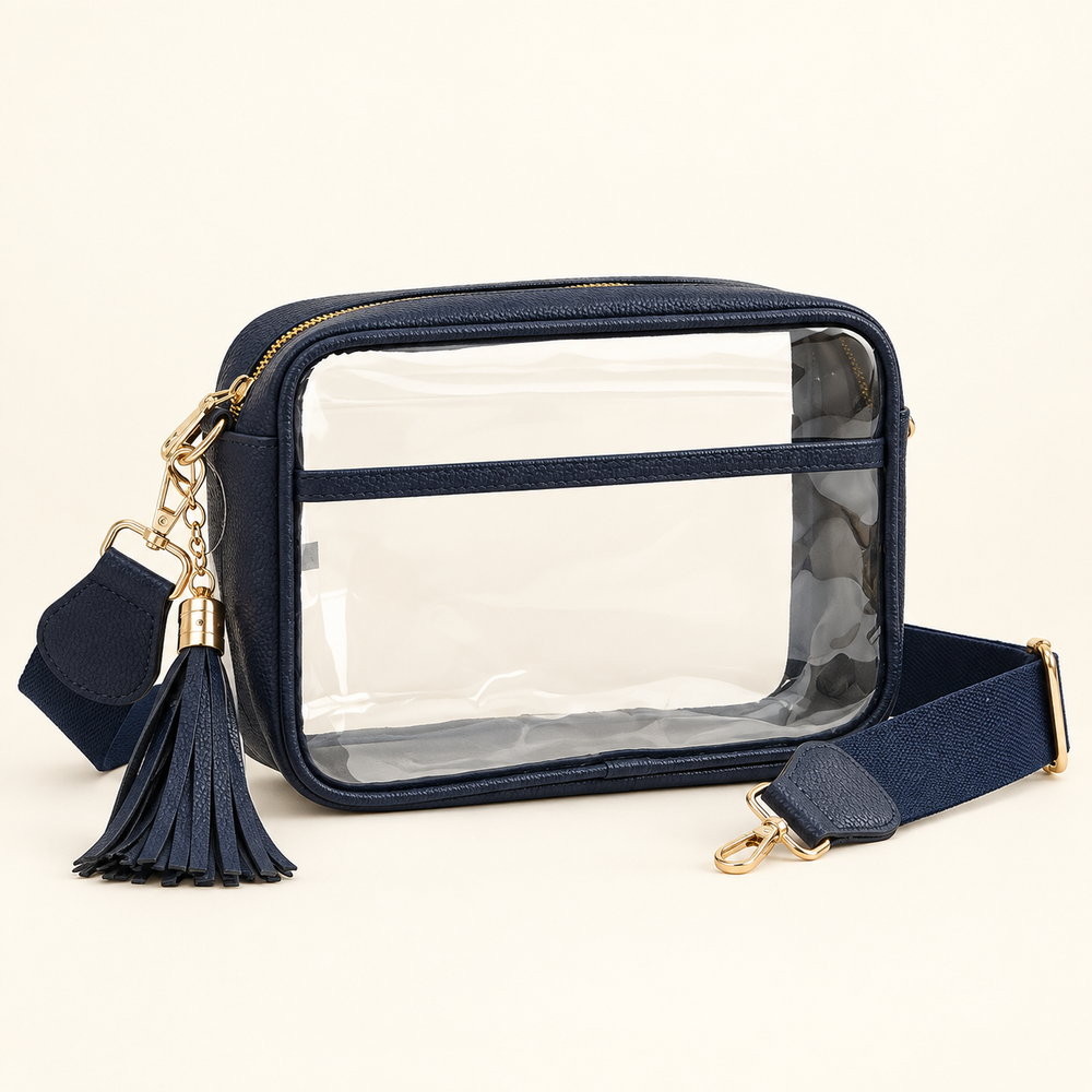 Clear stadium crossbody bag with navy trim, matching tassel and woven strap, and gold-tone hardware on a warm cream background.
