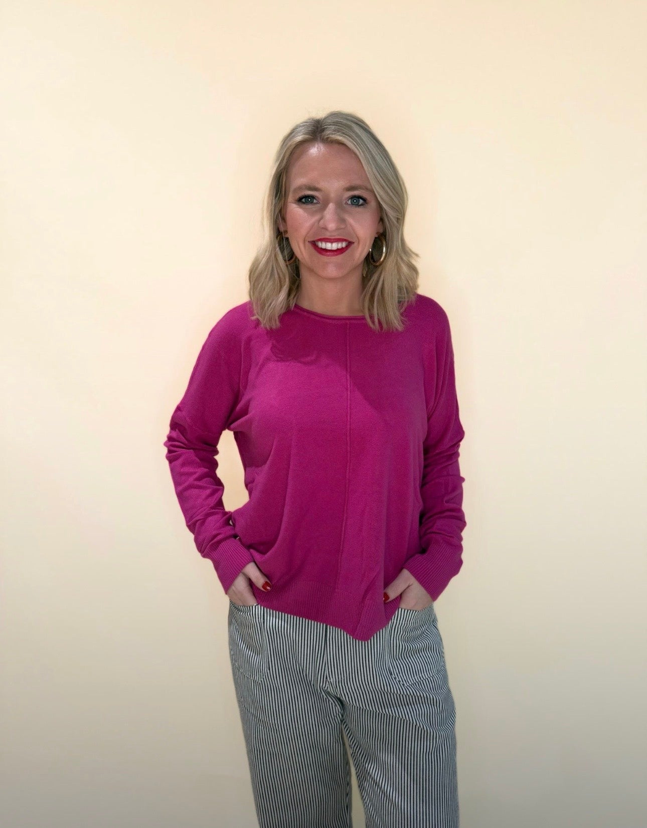 Front view of a woman wearing a fuchsia long-sleeve sweater paired with black and white striped pants, smiling with hands in pockets against a neutral cream background.