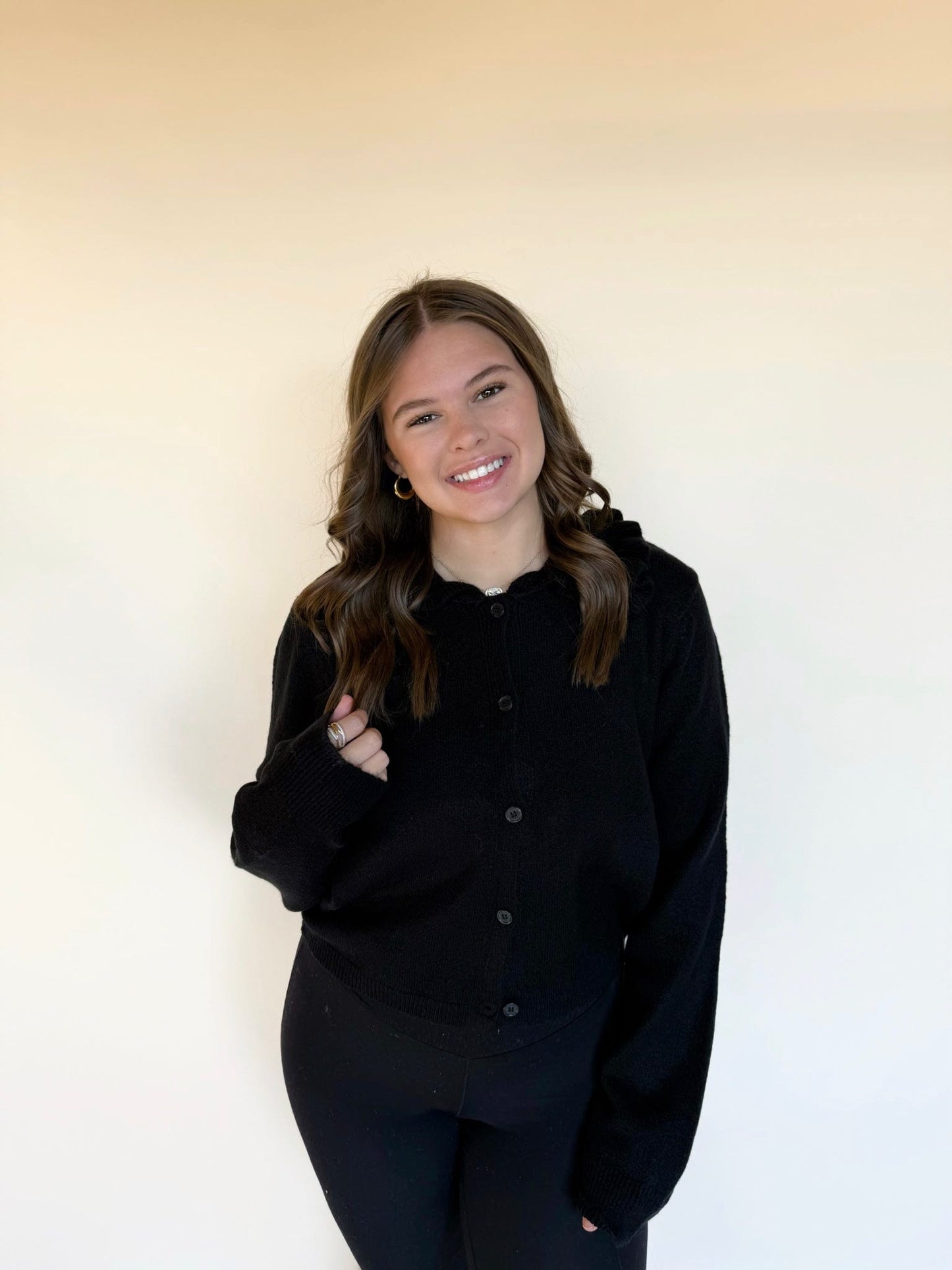Woman wearing a black button-front knit cardigan with long sleeves and black leggings, smiling in front of a light neutral background.