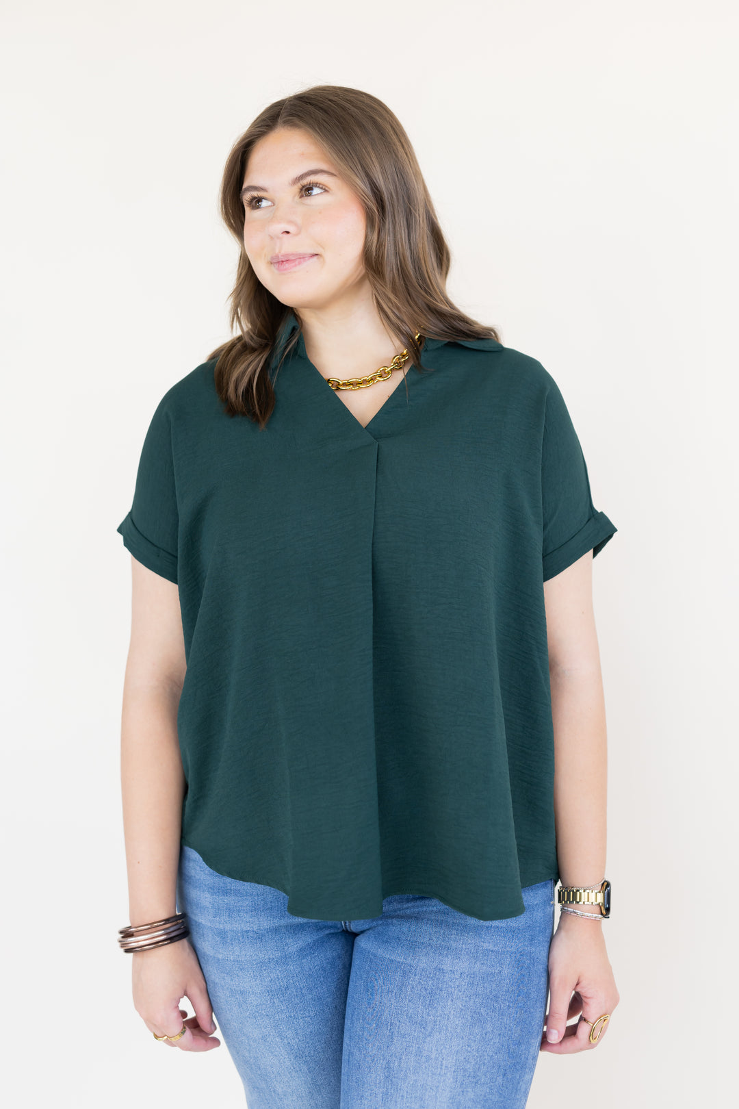 A woman wearing a dark green short-sleeve V-neck top and blue jeans, standing against a plain white background.