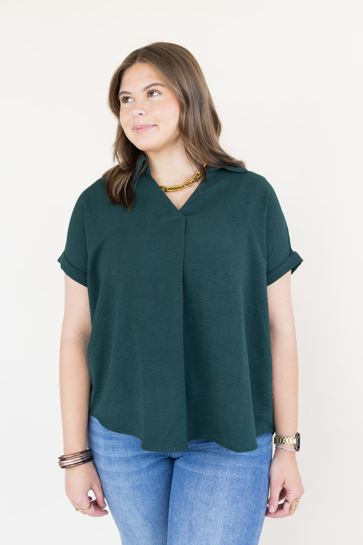 A woman wearing a dark green short-sleeve V-neck top and blue jeans, standing against a plain white background.