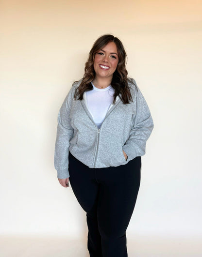 Woman wearing a light gray zip-up hoodie layered over a white top, posing casually with one hand in her pocket. 