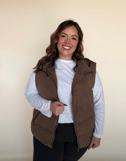 Woman wearing a chocolate brown puffer vest over a white long-sleeve top and black leggings, gently holding the zipper and smiling toward the camera.