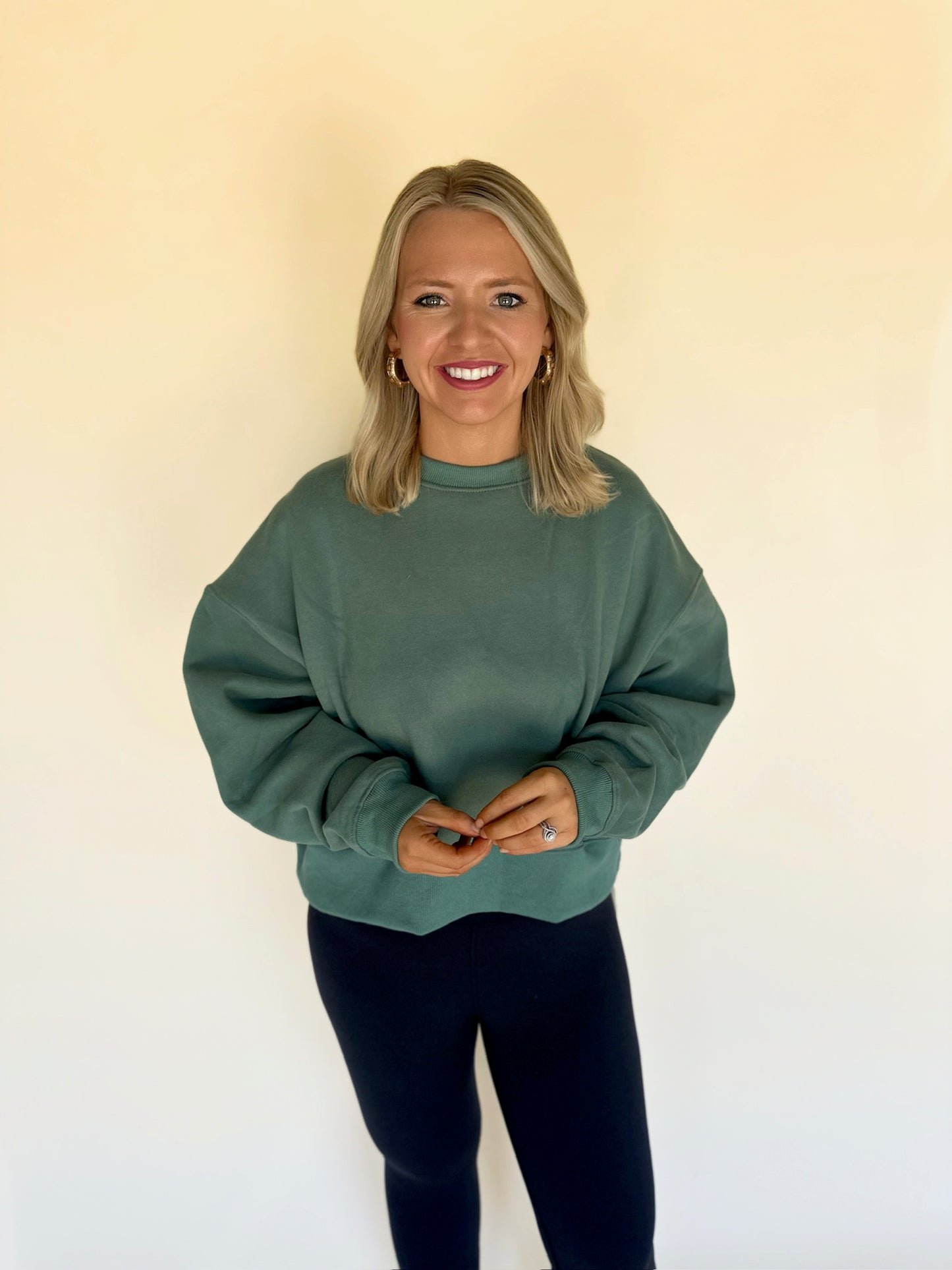 Woman wearing a green sweater and black pants against a beige wall