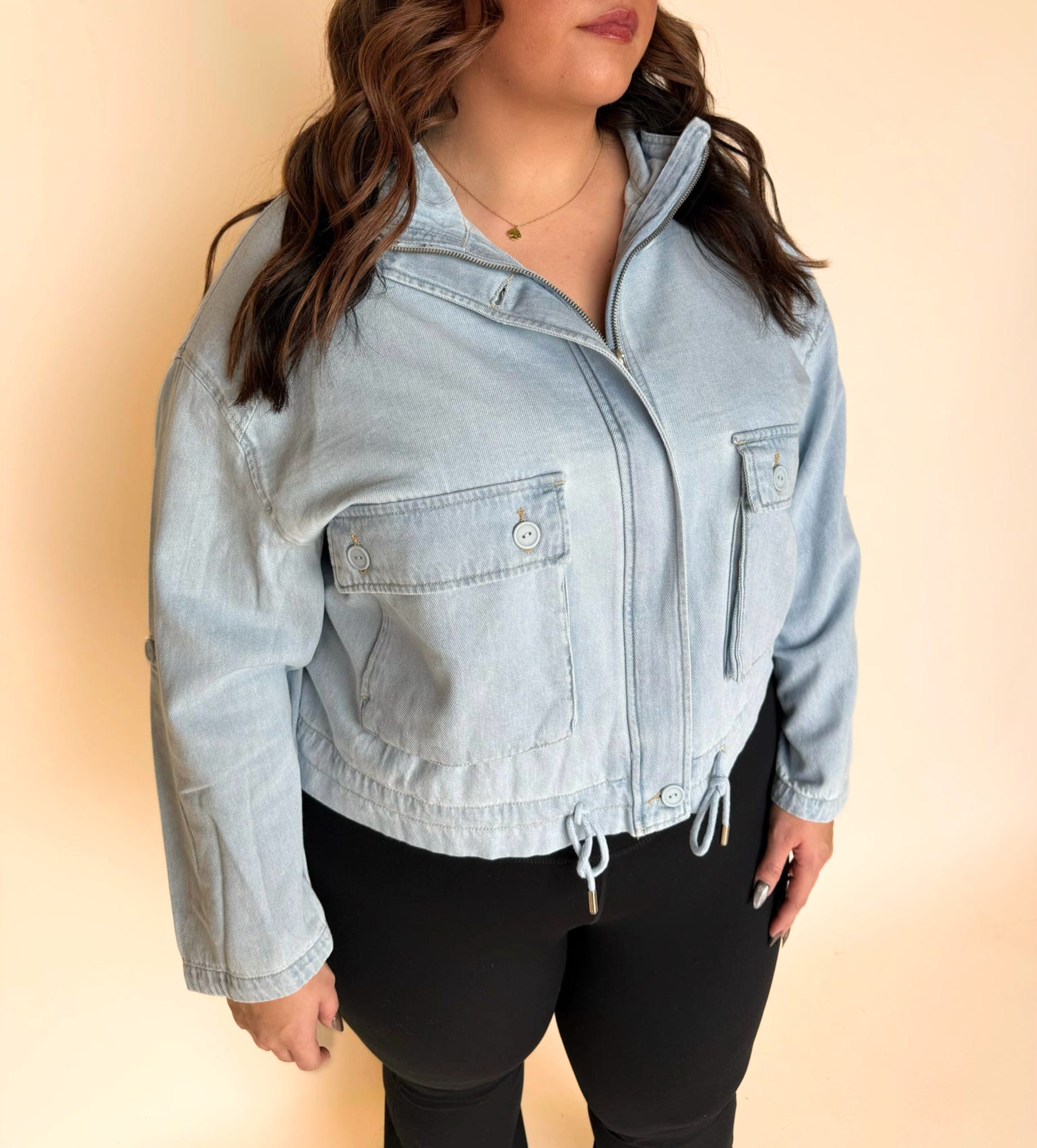 Close-up of a light-wash denim jacket featuring buttoned chest pockets, a zip front, and a drawstring hem, styled with black leggings in front of a neutral backdrop.