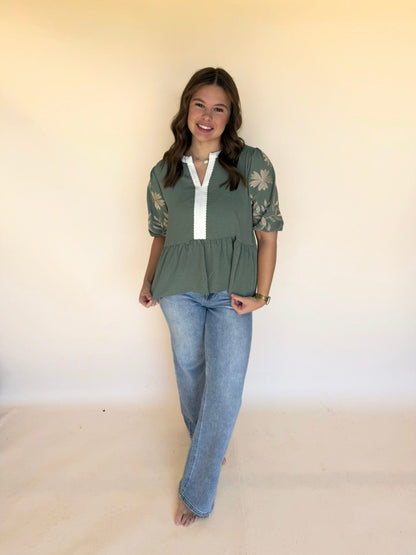 Woman smiling while wearing a sage green peplum-style embroidered blouse and light wash jeans, standing against a neutral background.