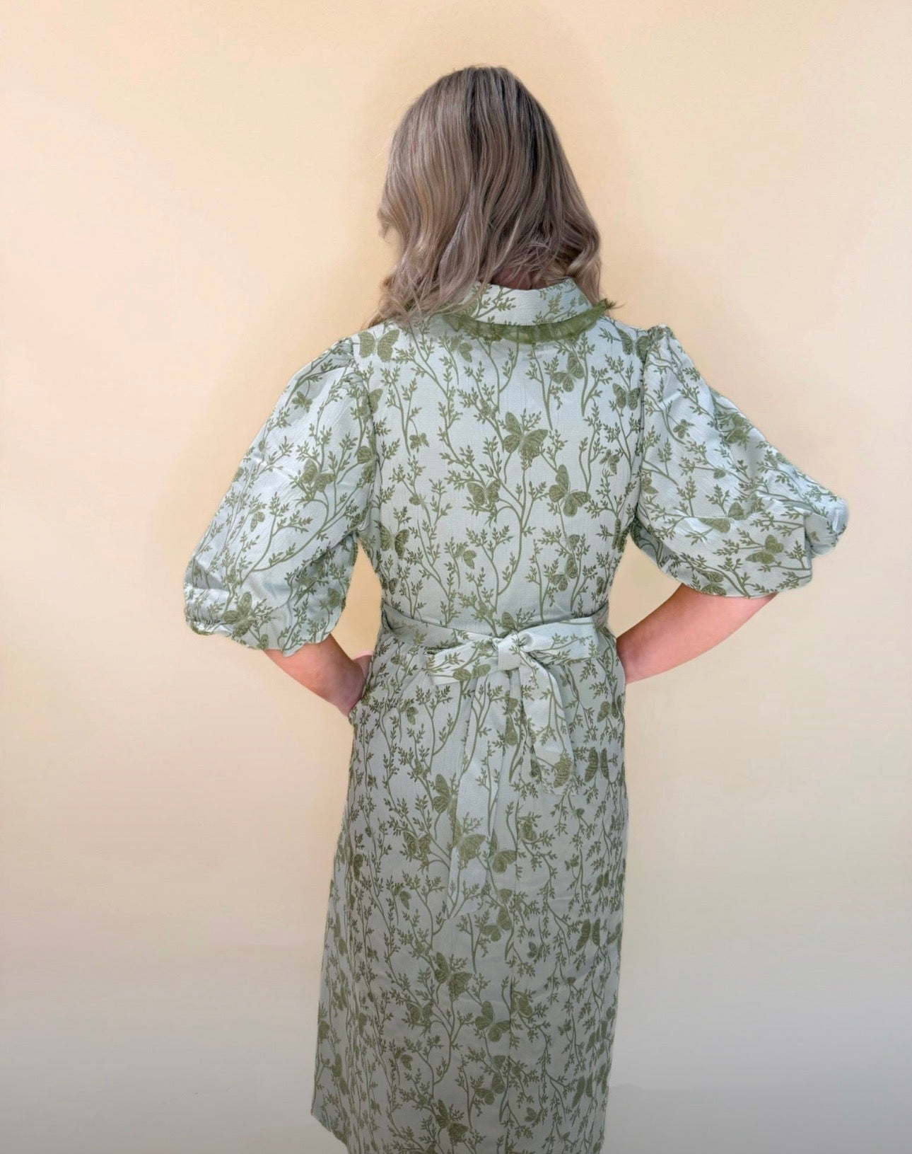 Back view of green floral print midi dress with puff sleeves and tie belt, showing full skirt flow and lightweight fabric against a cream backdrop.