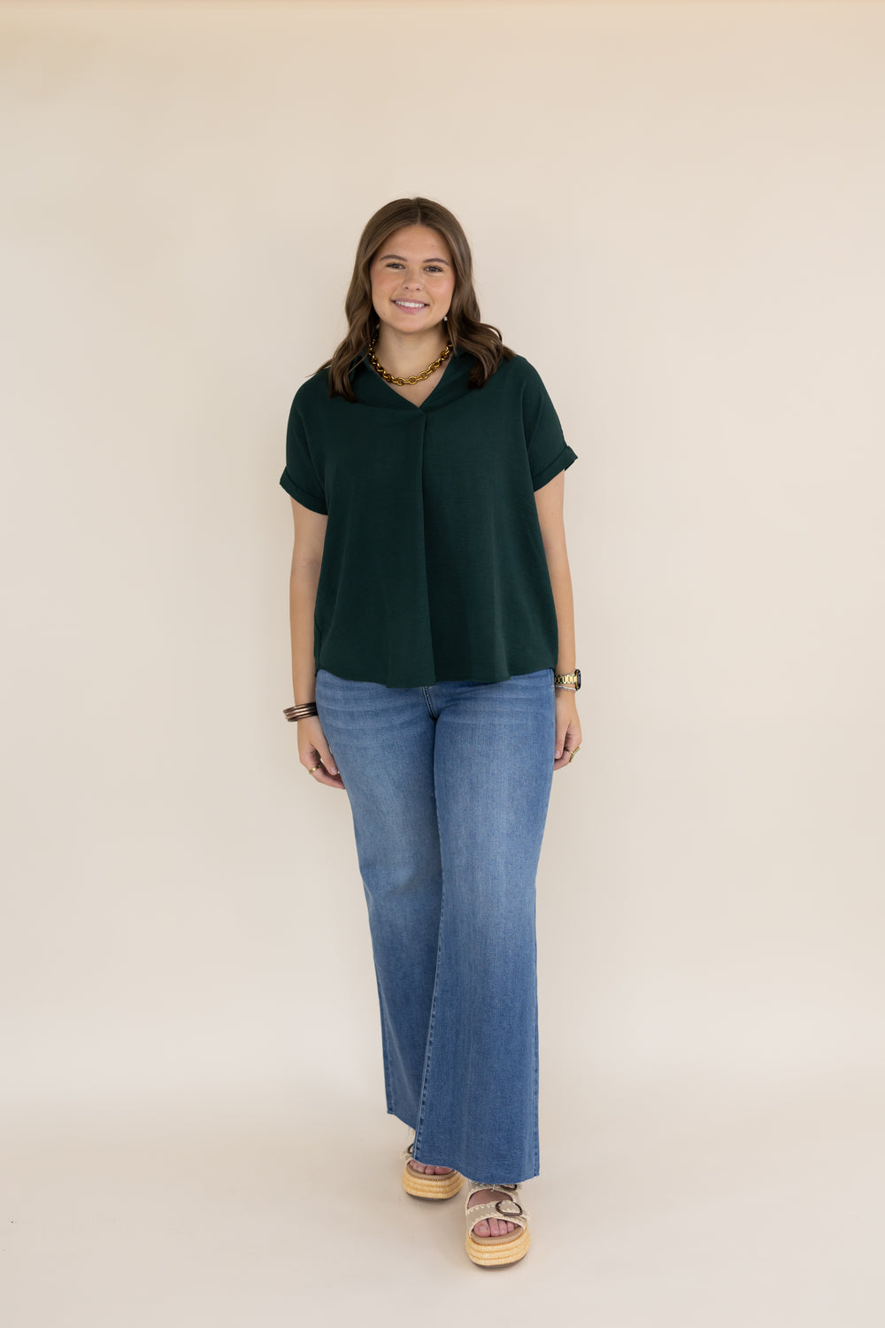 A woman modeling a dark green short-sleeve collared top with wide-leg blue jeans and patterned sandals, standing against a plain beige studio background.