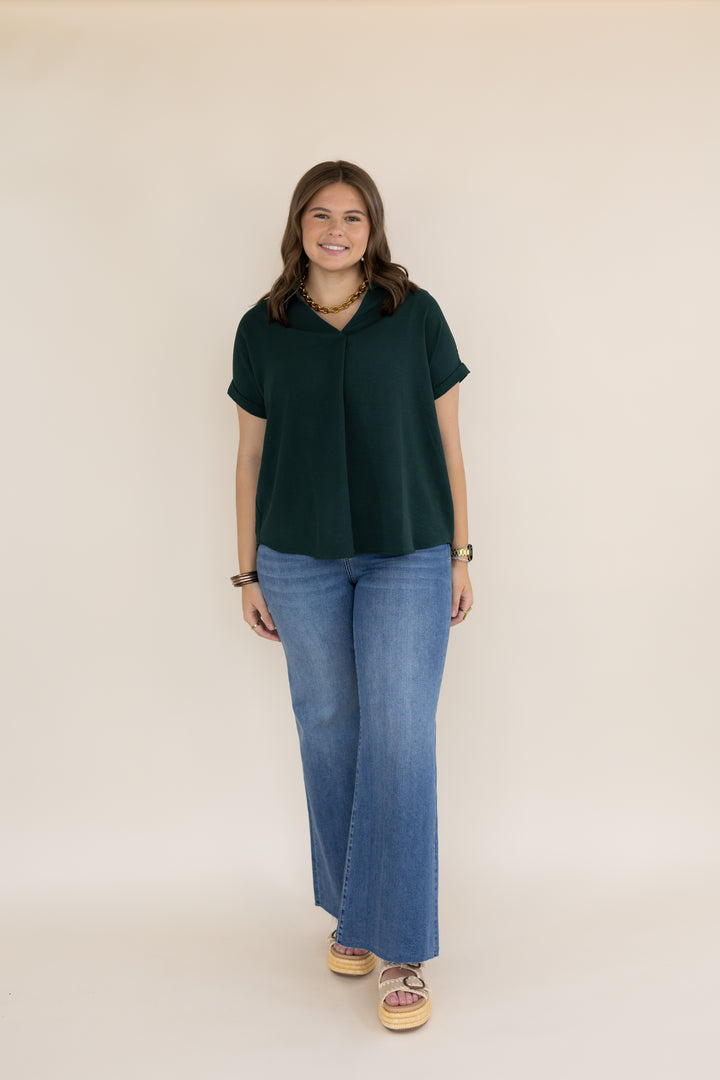 A woman modeling a dark green short-sleeve collared top with wide-leg blue jeans and patterned sandals, standing against a plain beige studio background.