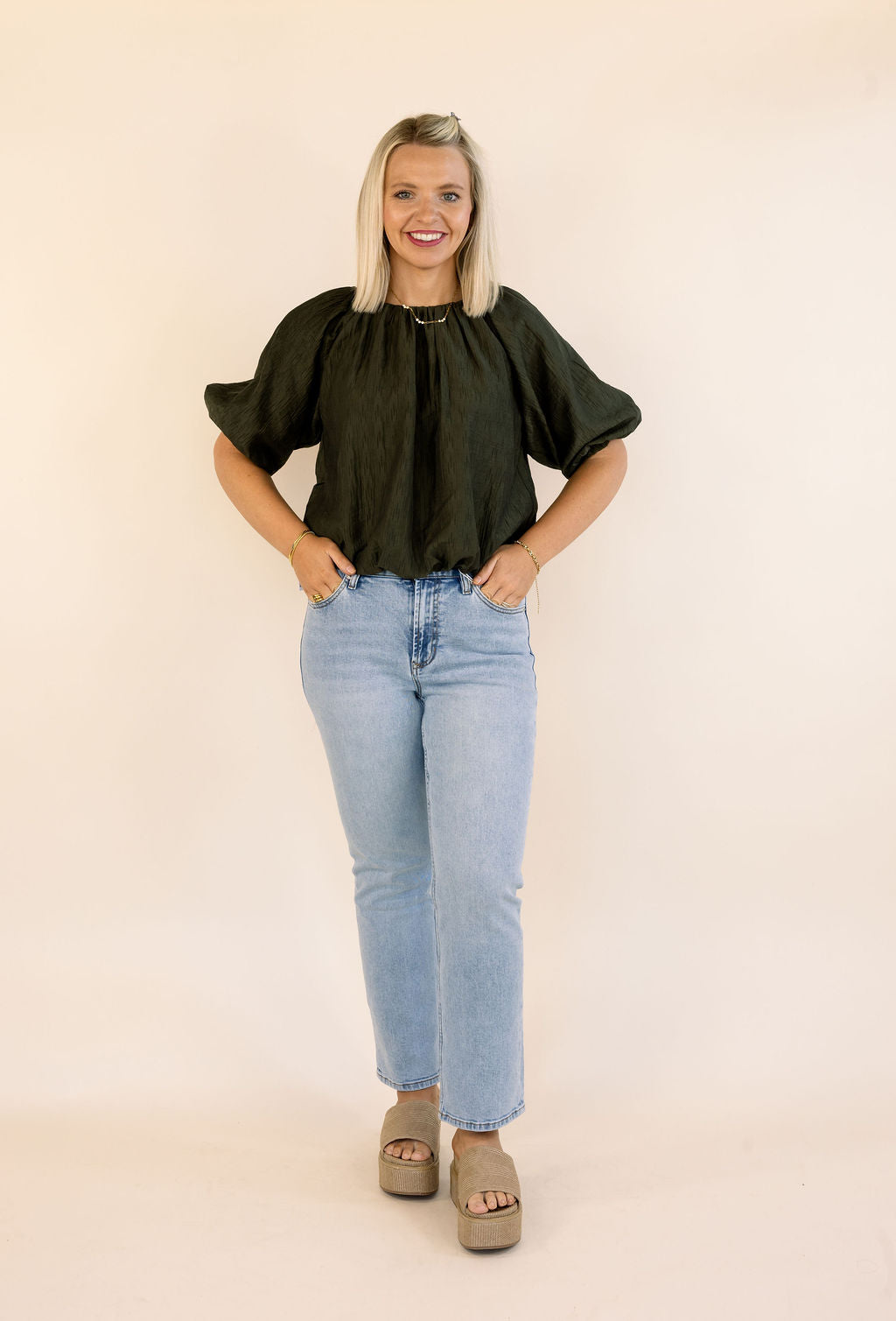 A woman modeling a dark olive puff-sleeve blouse with light-wash blue jeans and taupe platform sandals, standing against a solid cream backdrop.