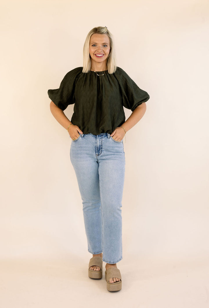A woman modeling a dark olive puff-sleeve blouse with light-wash blue jeans and taupe platform sandals, standing against a solid cream backdrop.