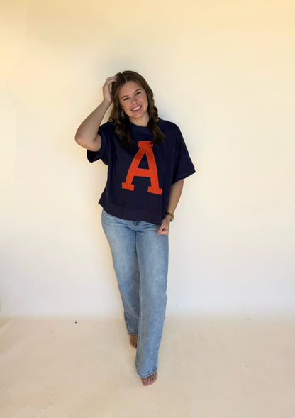Person wearing a navy blue t-shirt with a large orange 'A' and blue jeans against a plain background