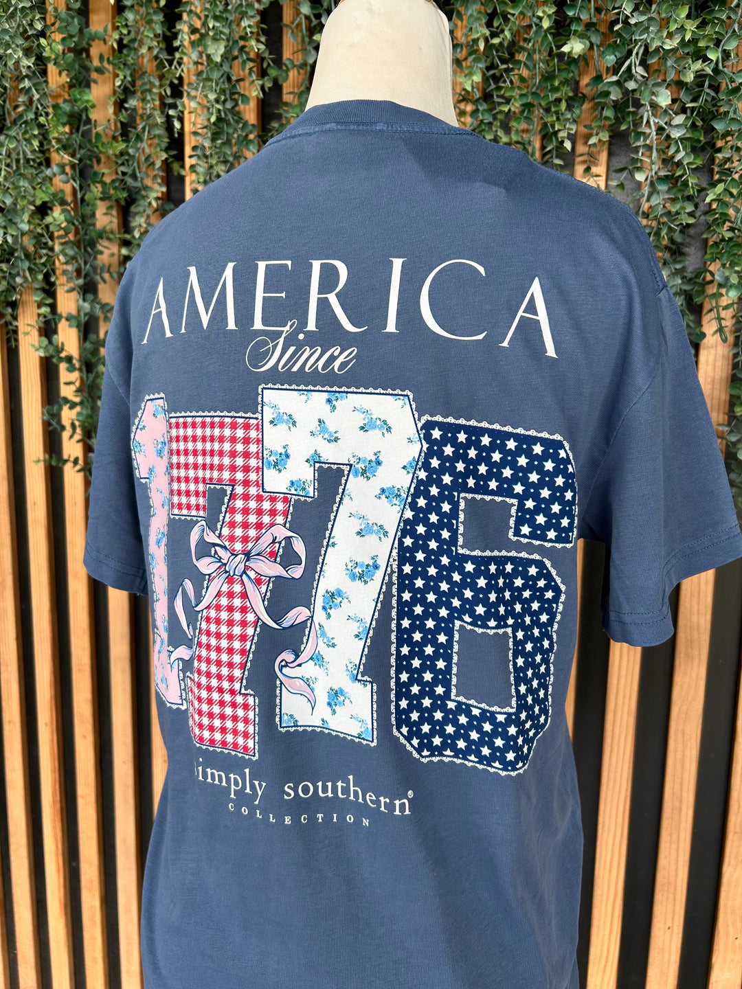 Back of a Simply Southern navy blue short sleeve graphic Youth T-shirt featuring "America Since 1776" with patriotic patchwork numbers in floral, gingham, and star prints accented by a decorative bow.
