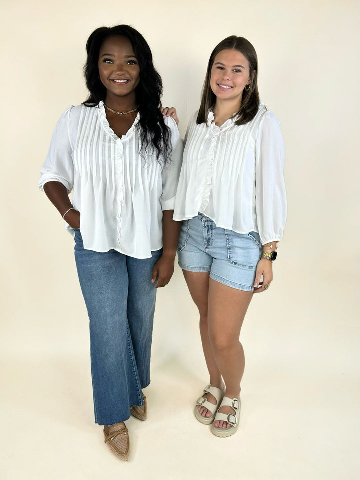 Woman wearing a white ruffle trim pintuck blouse with blue jeans standing beside another model in denim shorts.