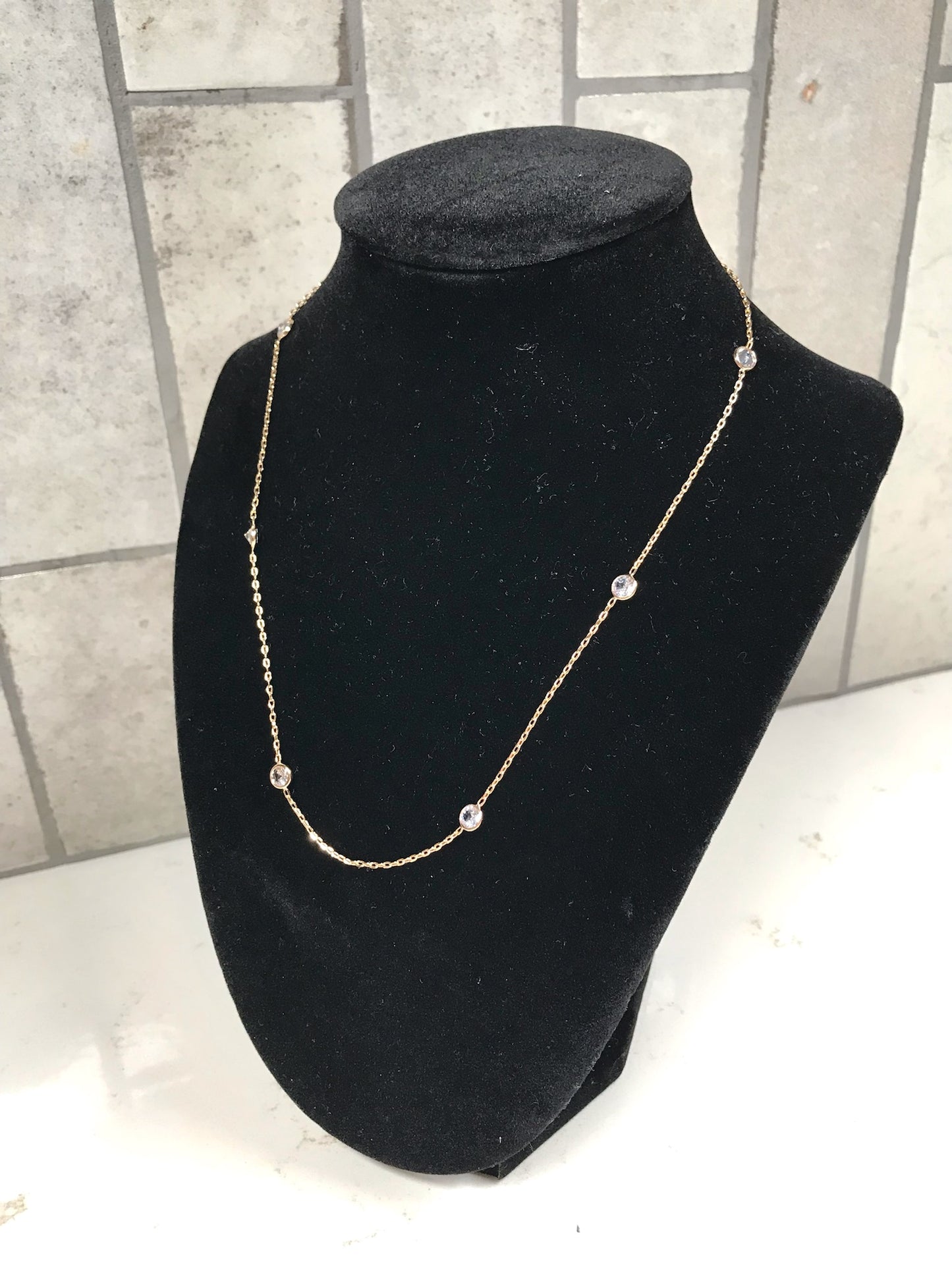 Gold dainty station necklace with small bezel-set clear crystals displayed on a black velvet necklace stand against a light tile background.