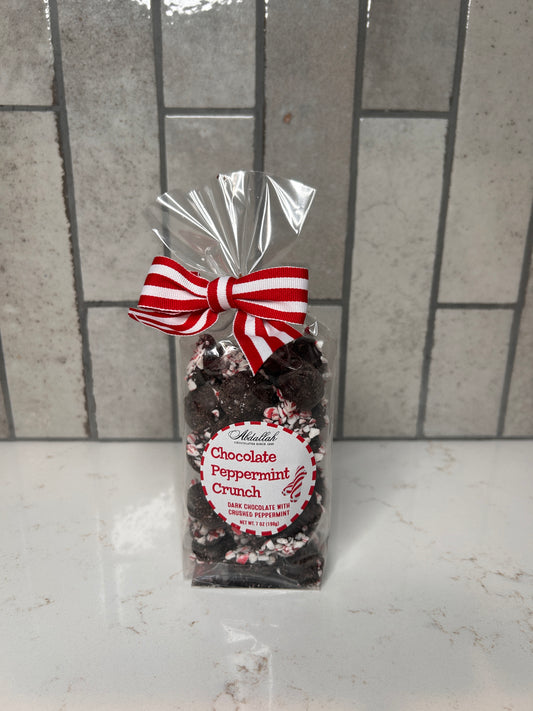 Clear bag of dark chocolate peppermint crunch candies coated with crushed peppermint, tied with a red-and-white striped bow, displayed on a light countertop with a neutral tile backsplash.