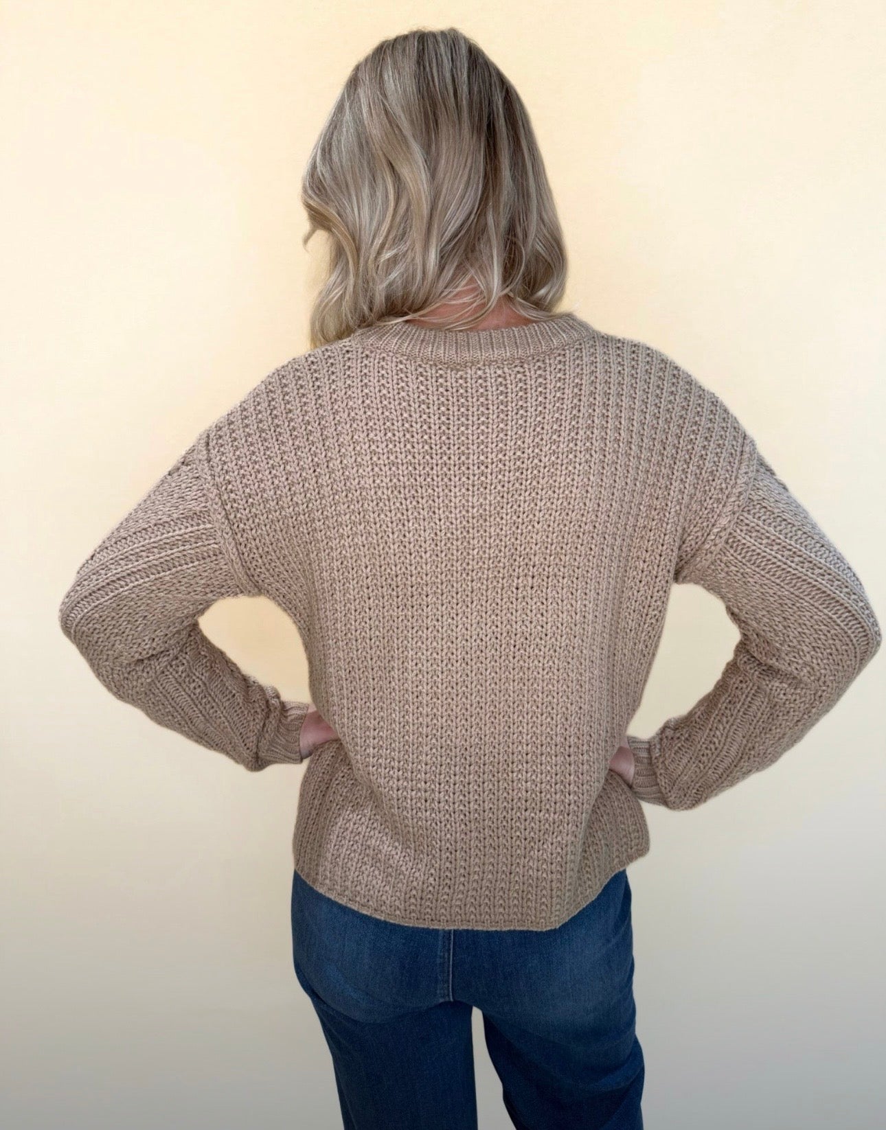 Back view of tan cable knit sweater showing ribbed knit texture and relaxed fit, paired with blue jeans against a neutral background.