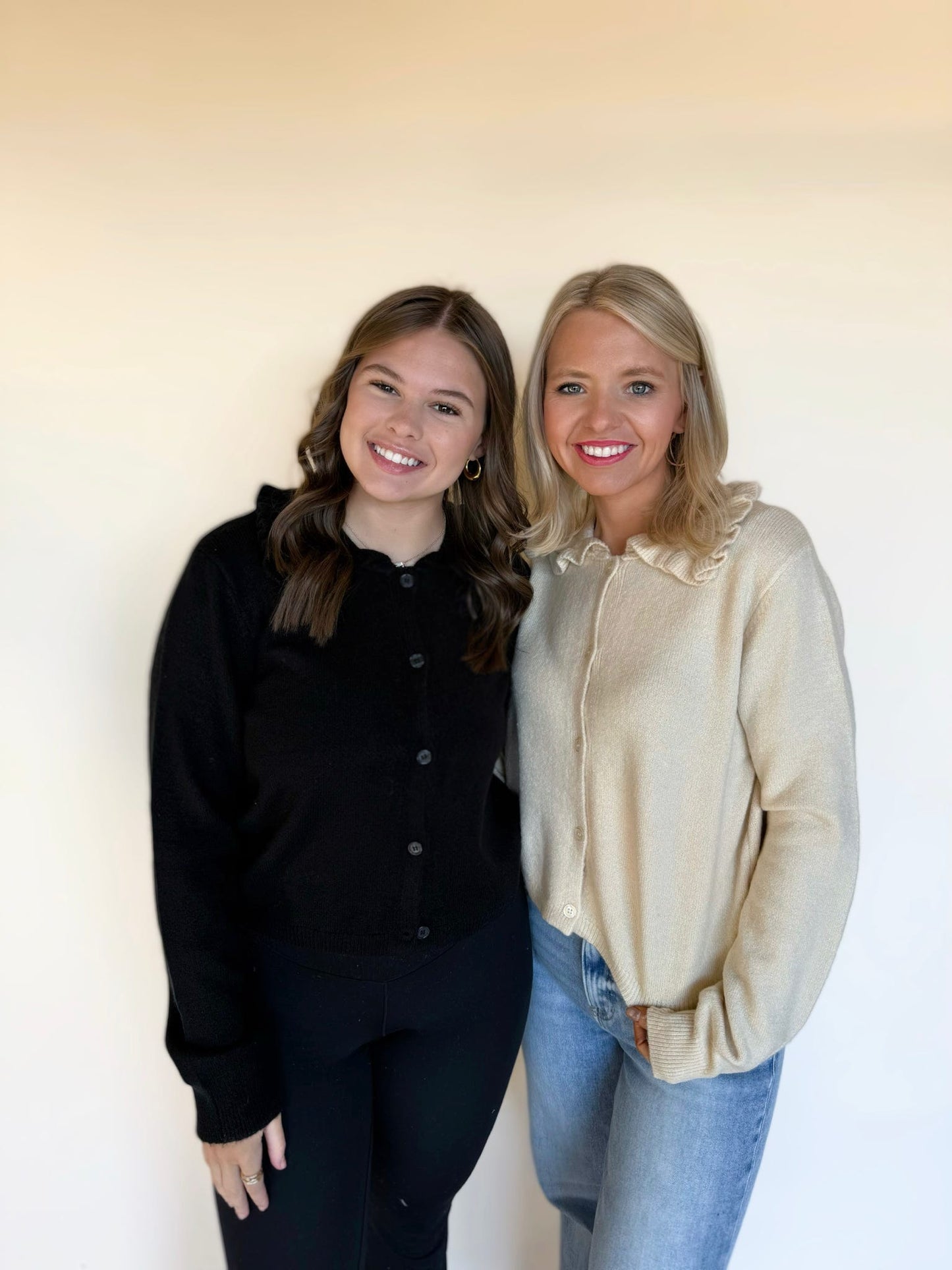 Two women standing side by side wearing knit button-front cardigans; one in a cream ruffle-collar sweater with blue jeans and the other in a black cardigan with black leggings, smiling in front of a neutral background.
