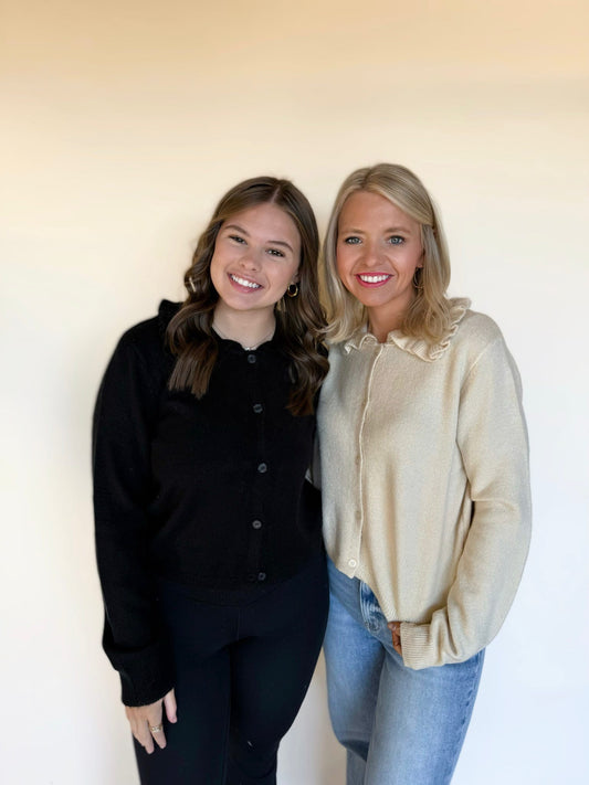 Two women standing side by side wearing knit button-front cardigans; one in a cream ruffle-collar sweater with blue jeans and the other in a black cardigan with black leggings, smiling in front of a neutral background.