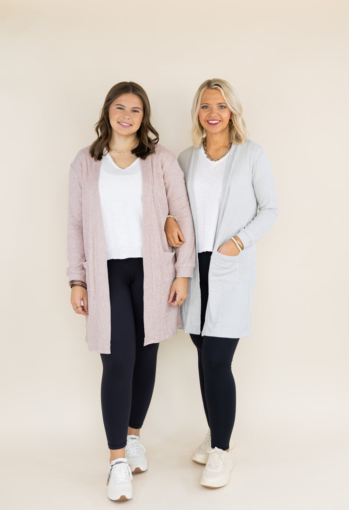 Two women standing side by side wearing matching long open-front cardigans over white tops and black leggings, photographed against a plain warm beige studio background.