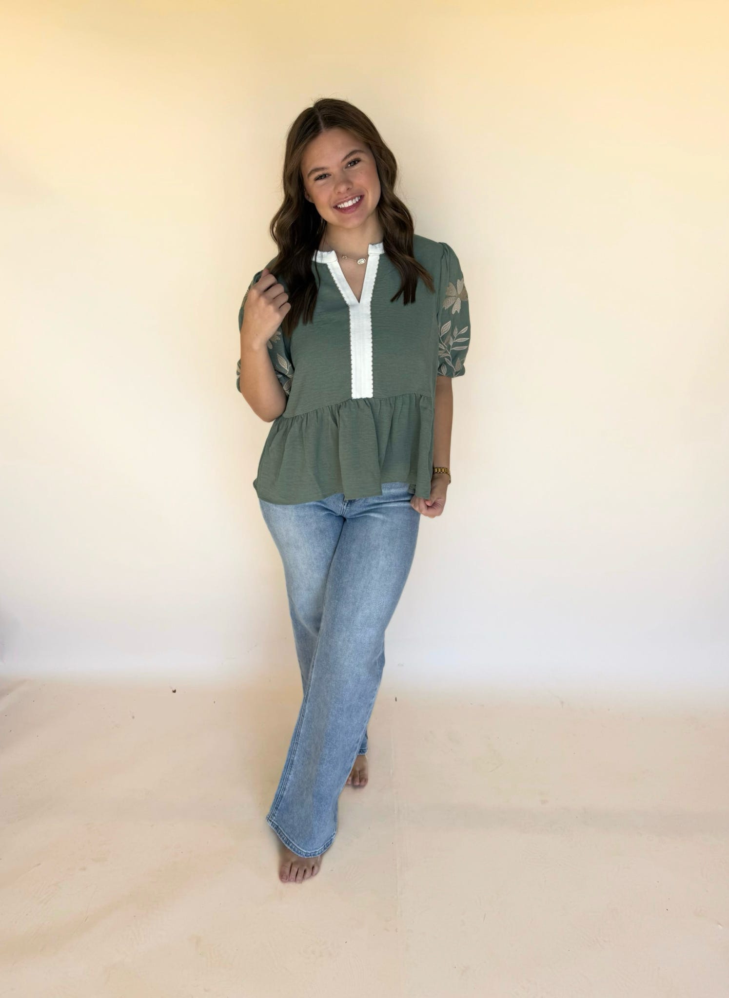 Woman posing in a sage green blouse with floral embroidered sleeves and a white split neckline, paired with light wash jeans.
