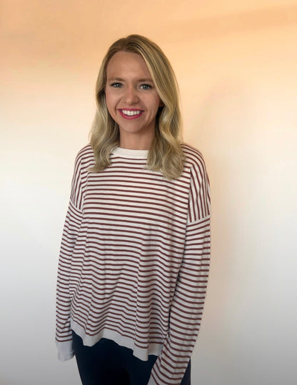 Woman in a cream and rust striped long-sleeve sweater with black pants, smiling and standing against a neutral beige background. Casual cozy fall top.