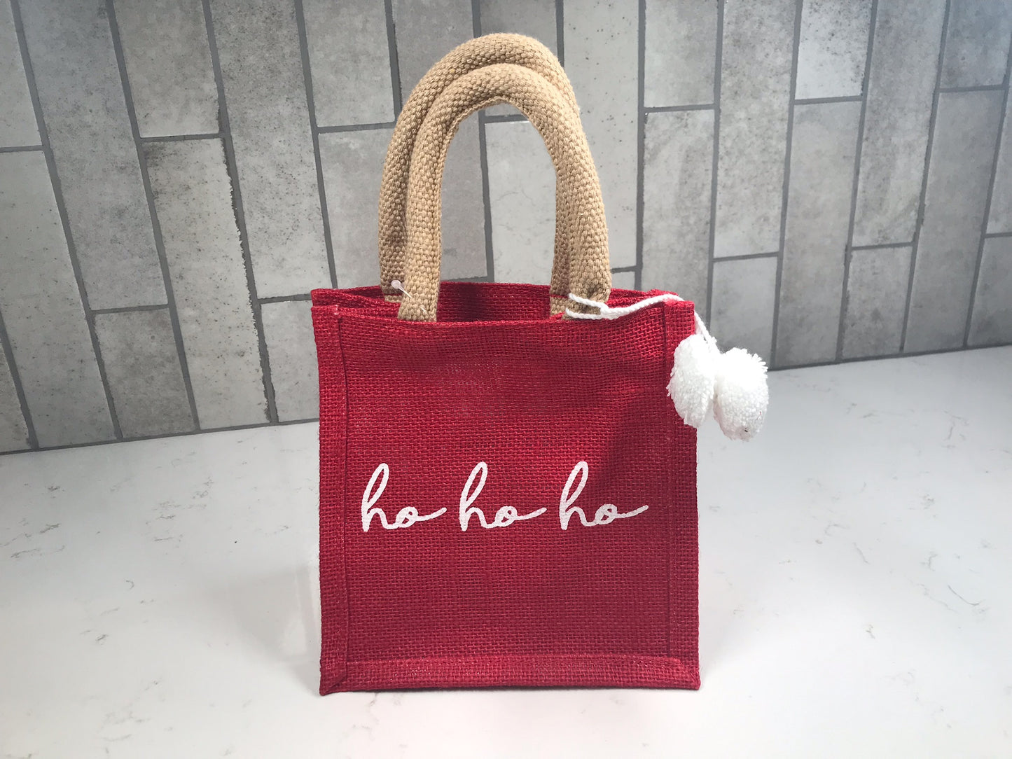 Red petite gift tote with “ho ho ho” script, natural rope handles, and white pom accents, displayed upright on a light tile background.