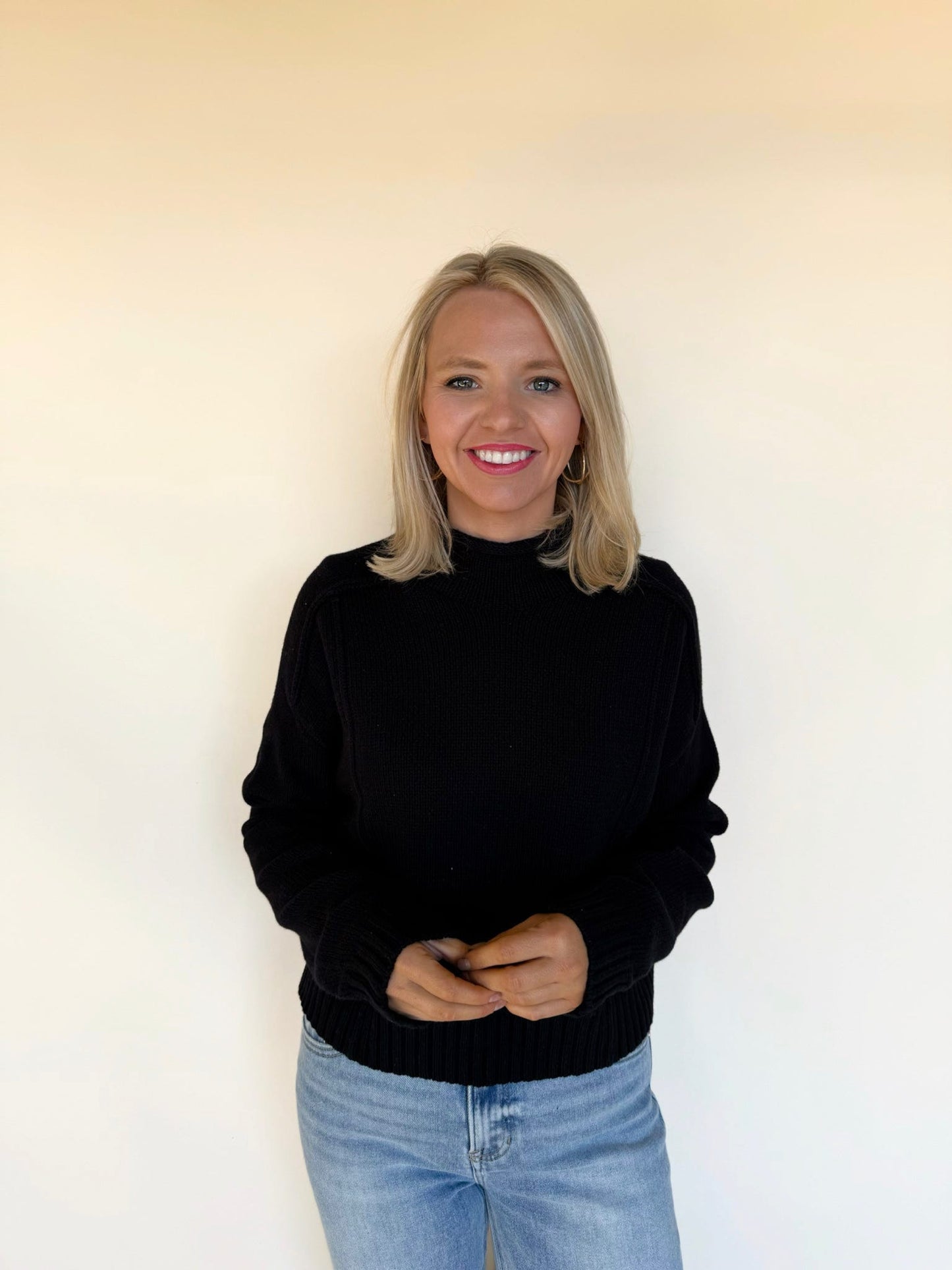 Woman wearing a black mock-neck knit sweater with long sleeves and ribbed details, paired with light-wash jeans in front of a neutral background.