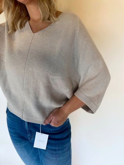 Person wearing an oatmeal sweater with a tag, standing against a plain background