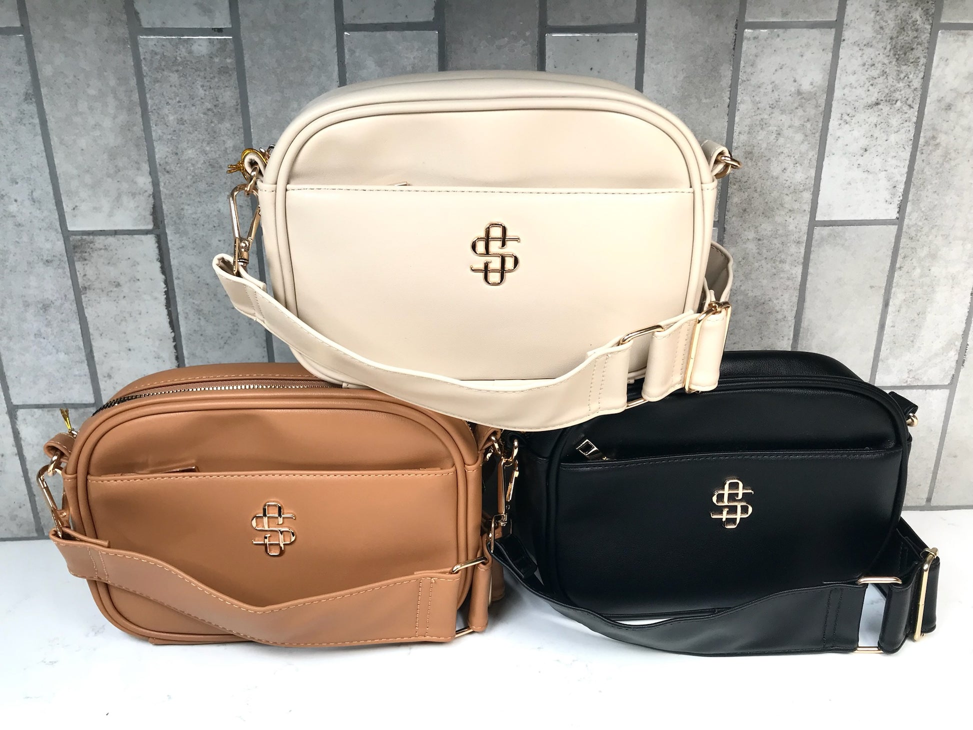 Three bags in beige, brown, and black with a visible brand logo on a gray tiled background.