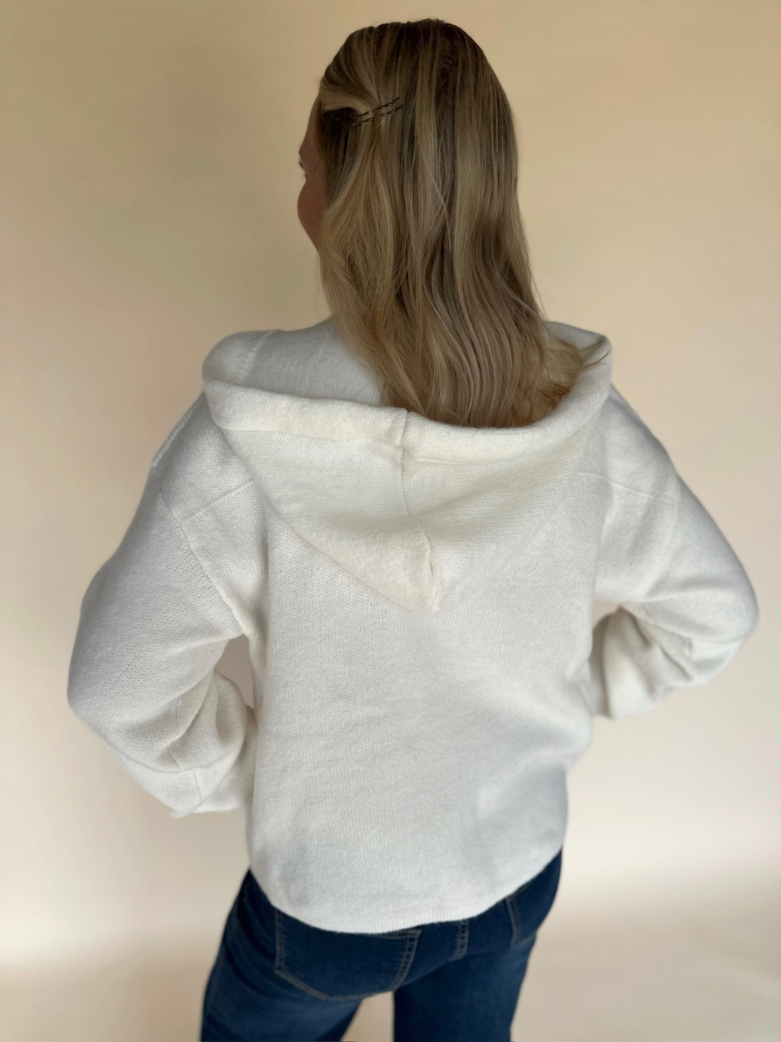Back view of a woman wearing a cream knit hoodie cardigan, showing the relaxed hood and soft knit texture, paired with dark denim jeans against a neutral backdrop.