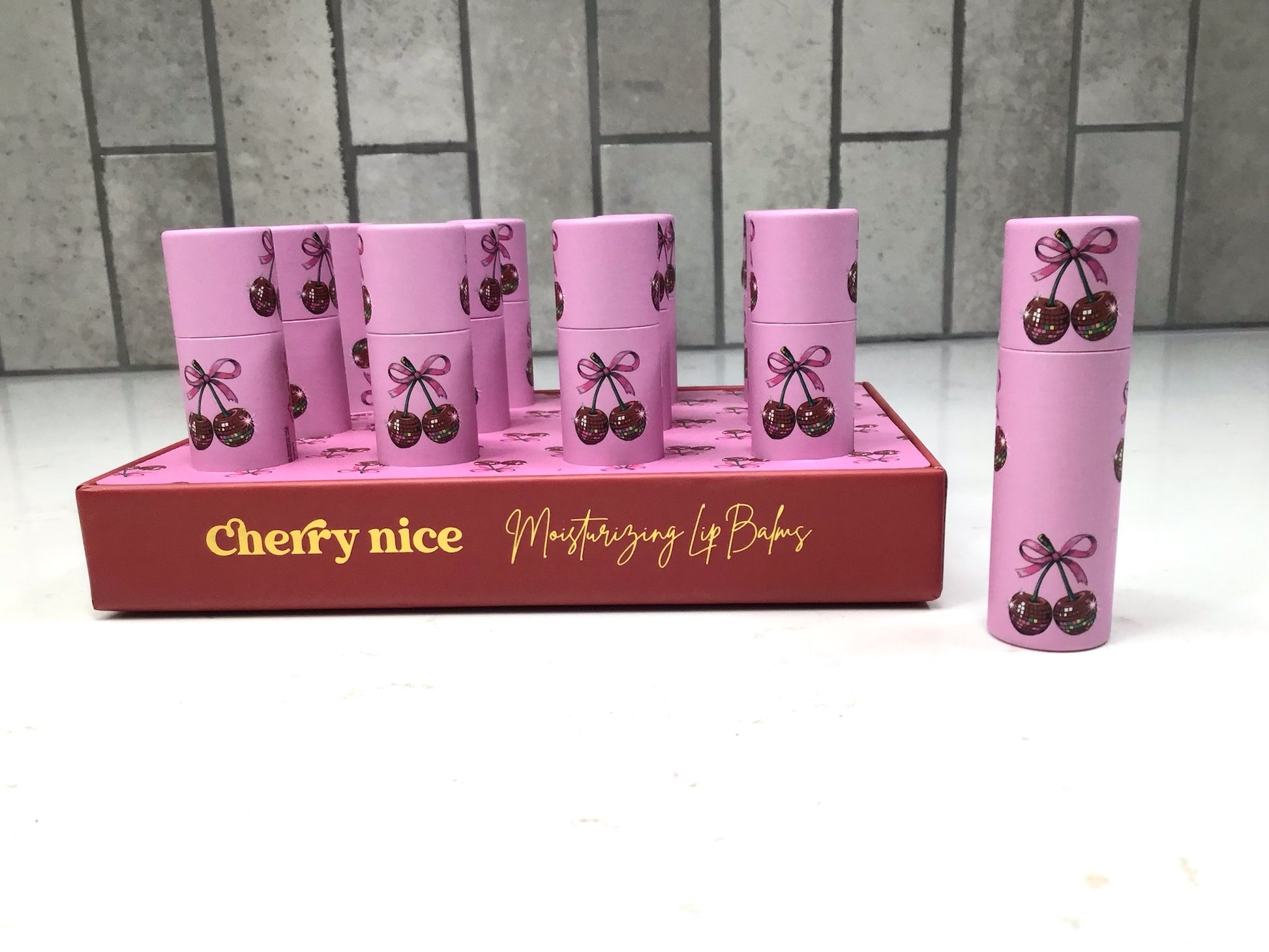 Set of pink lip balms with cherry design in a display box on a white surface.