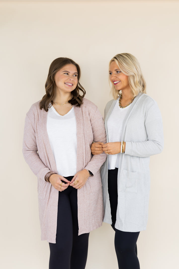Two women modeling long open-front cardigans in a studio setting. The woman on the left wears a light blush pink cardigan over a white top and black pants, while the woman on the right wears a light gray cardigan over a white top and black pants.