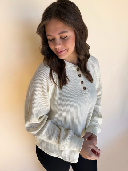 Woman wearing a cream ribbed sweater with button details against a plain background