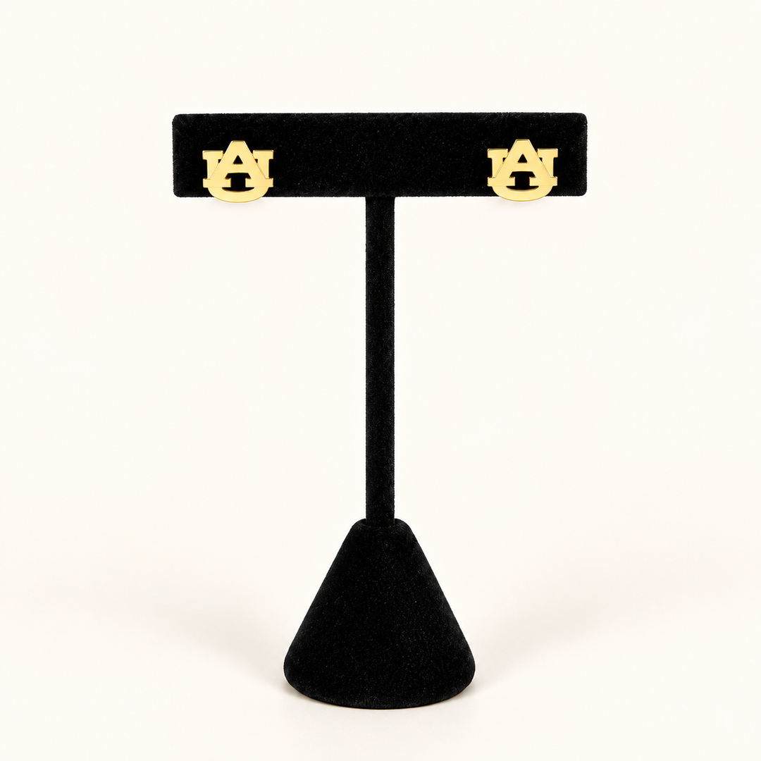 Gold Auburn interlocking AU stud earrings displayed on a black velvet jewelry stand against a warm cream background.