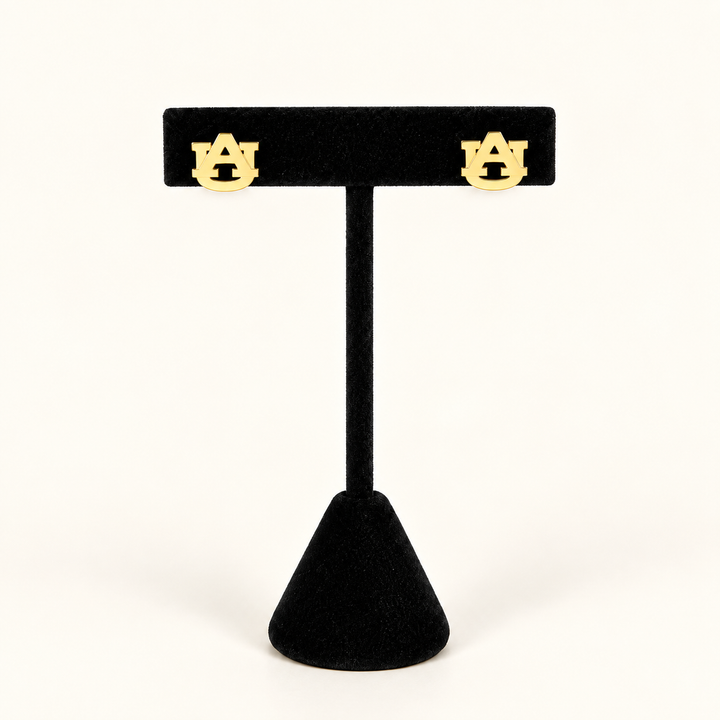 Gold Auburn interlocking AU stud earrings displayed on a black velvet jewelry stand against a warm cream background.