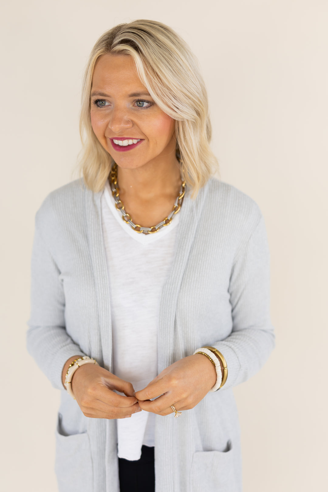 A blonde woman wearing a light gray ribbed cardigan over a white t-shirt, accessorized with a chunky gold chain necklace and gold bracelets, posing against a solid beige background.