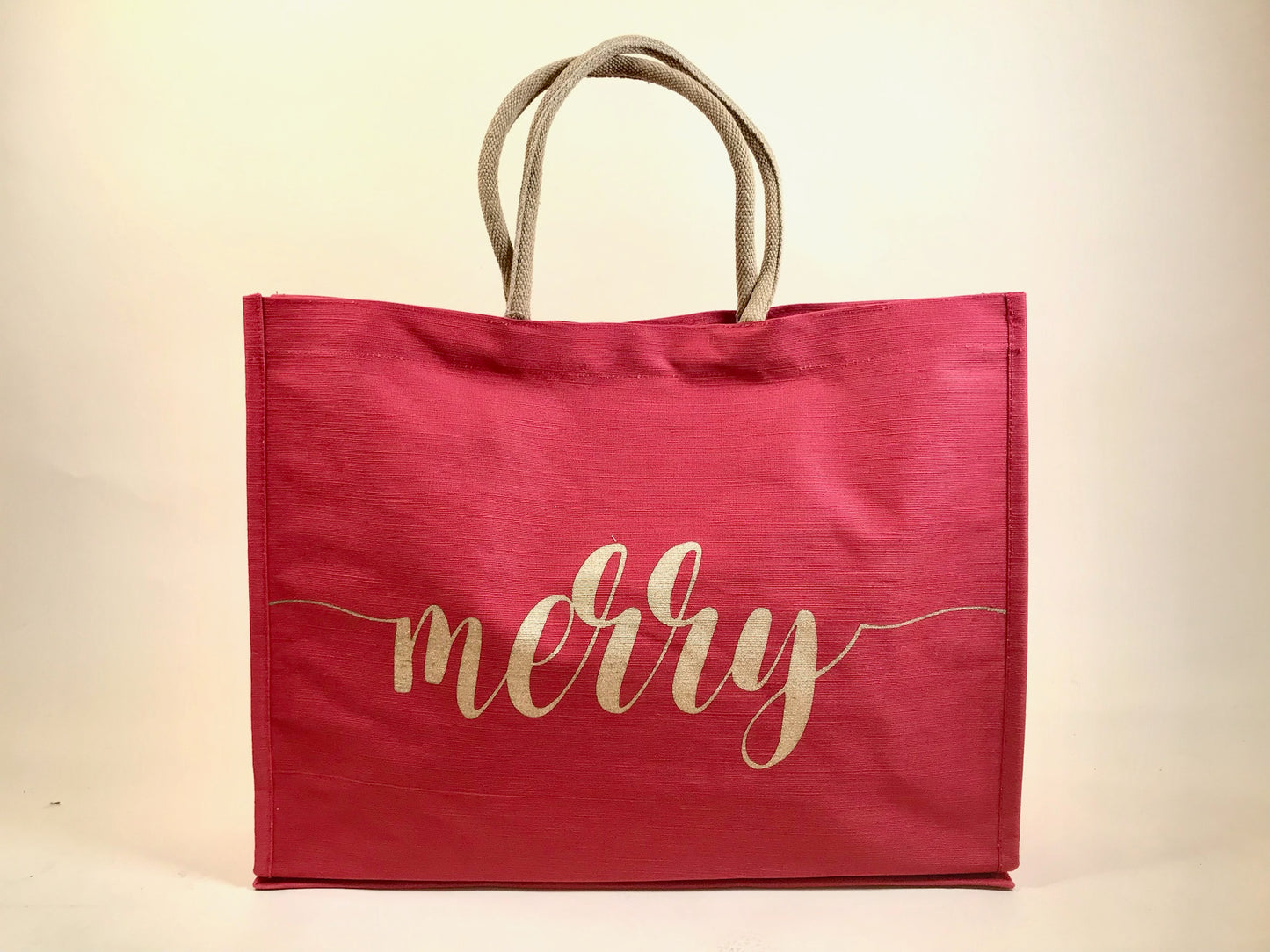 Large red holiday tote bag with gold “merry” script and rope handles against neutral backdrop