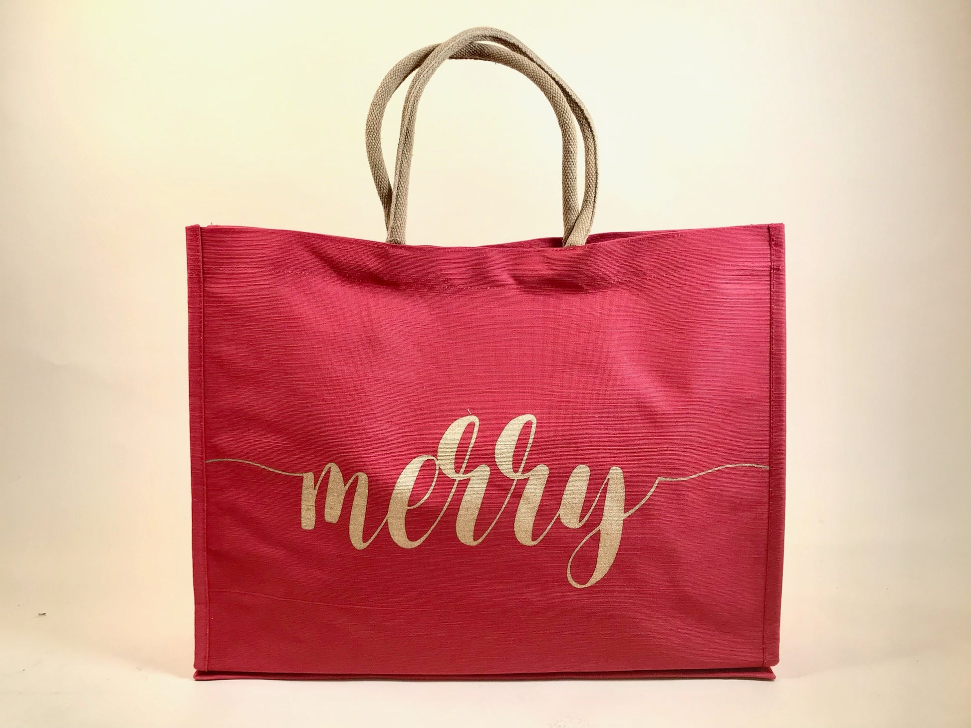 Large red holiday tote bag with gold “merry” script and rope handles against neutral backdrop
