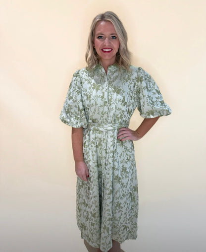 Full body front view of green floral puff sleeve midi dress with belted waist and button front, model smiling with one hand on hip against a soft cream background.