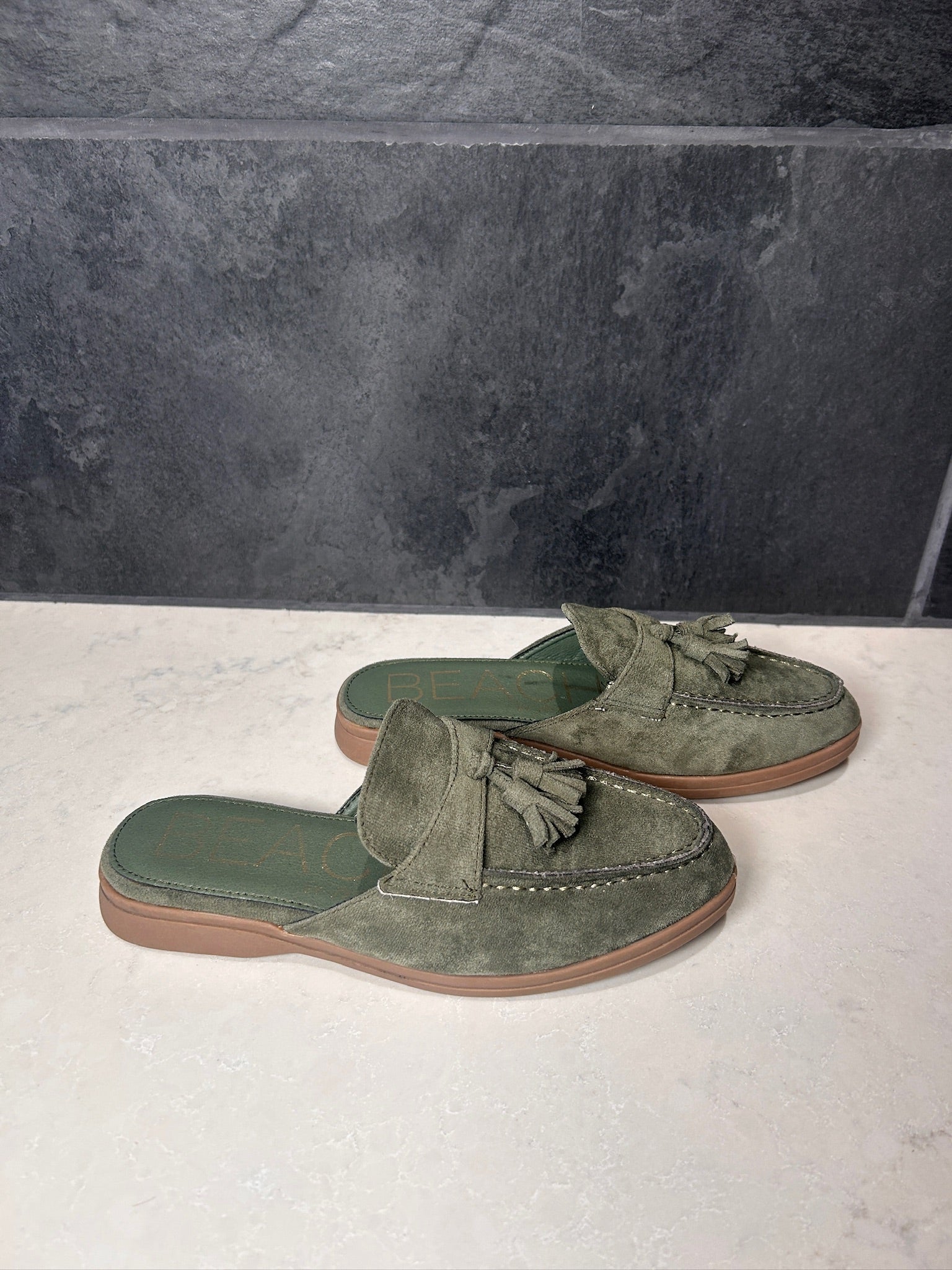 Pair of green suede loafers with tassels on a light surface with a dark tiled wall background.