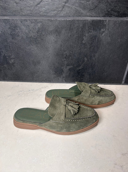 Pair of green suede loafers with tassels on a light surface with a dark tiled wall background.