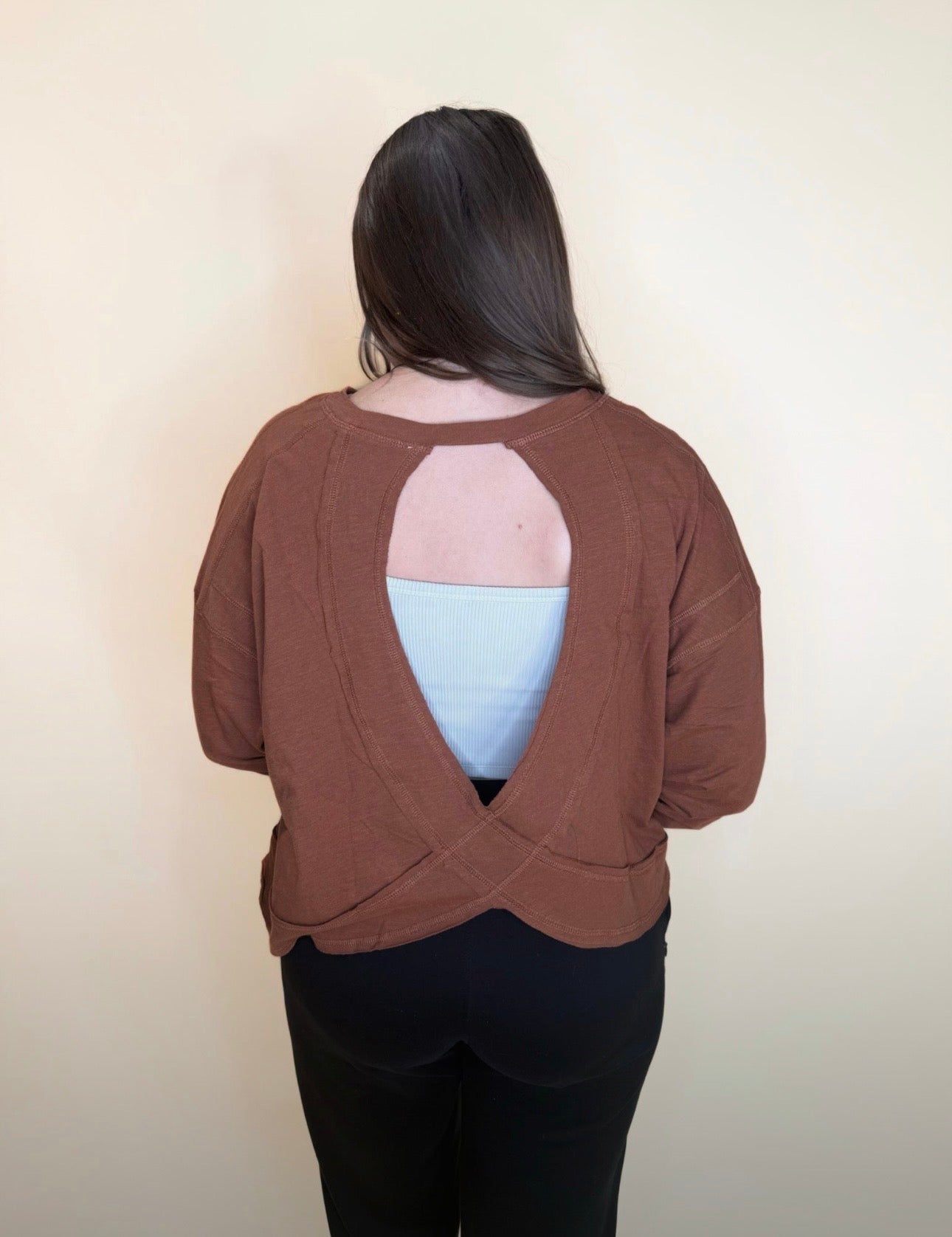Back view of a brown long sleeve top featuring an open cutout and criss-cross hem, layered over a light blue tank, paired with black pants for a casual outfit.