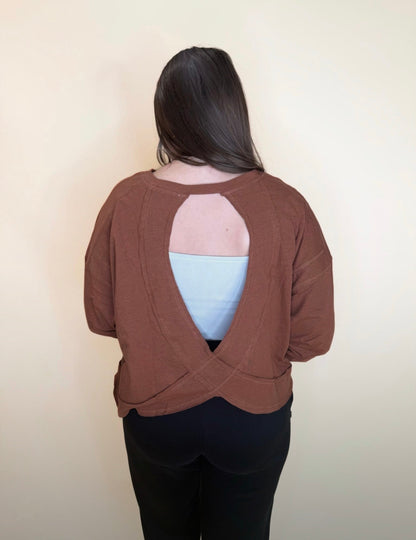 Back view of a brown long sleeve top featuring an open cutout and criss-cross hem, layered over a light blue tank, paired with black pants for a casual outfit.