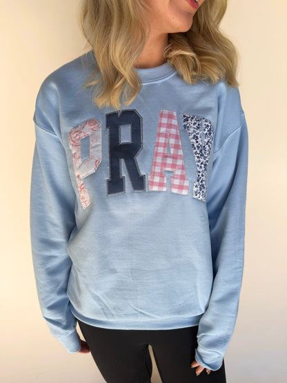 Close-up of a light blue sweatshirt with patchwork appliqué “PRAY” lettering in floral, gingham, and navy fabrics.