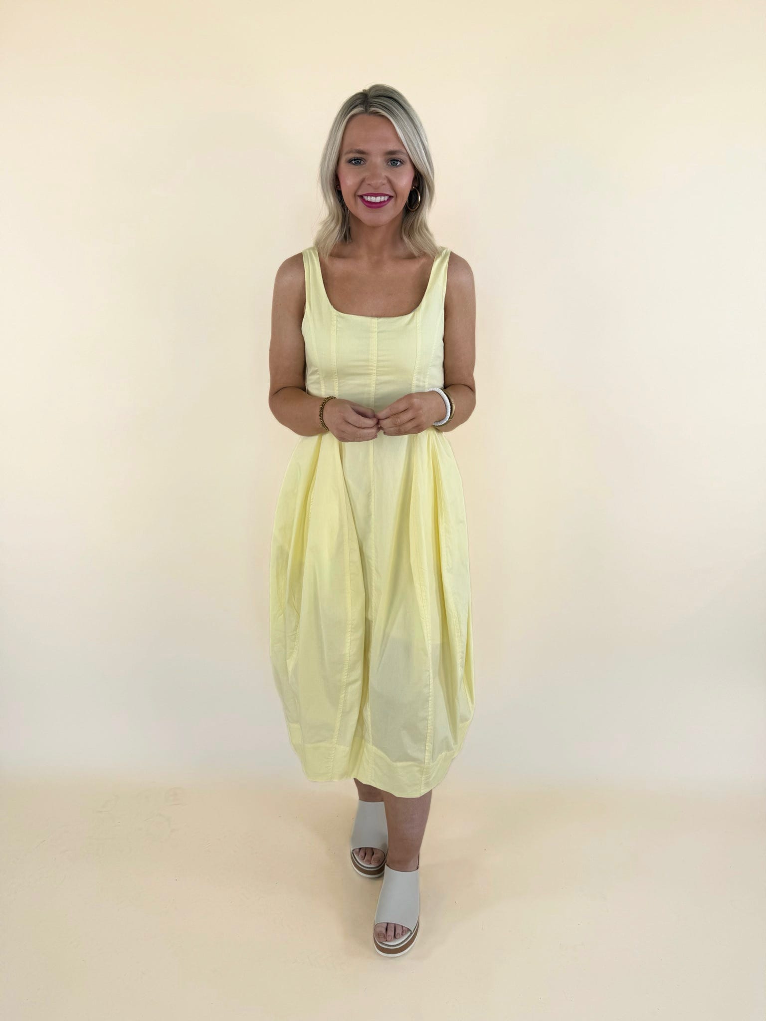 Woman wearing butter yellow midi dress with tulip hem, square neckline, and flowy silhouette styled with neutral slide sandals